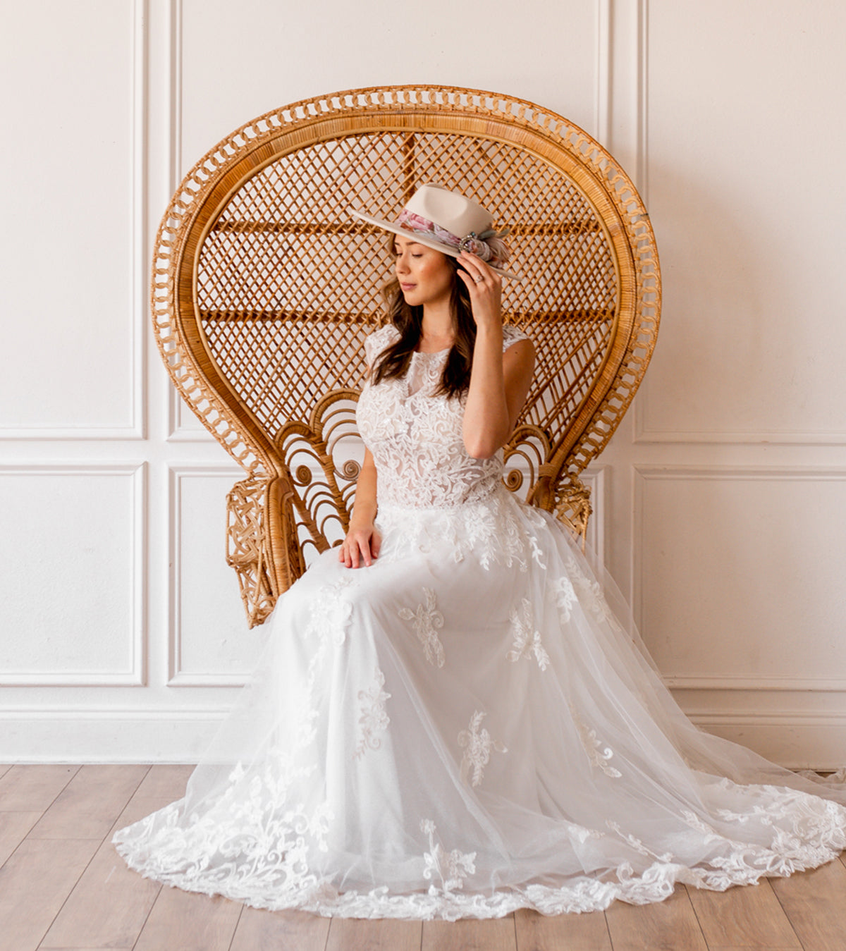 Bride sitting in a chair wearing a wedding dress and a white flat brim hat. 