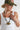A woman wearing an olive green embroidered butterfly trucker hat  paired with a white ribbed tank top and a black beaded necklace with a gold ginkgo leaf pendant-FLEASTYLE.