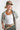 A woman wearing an olive green trucker cap with a butterfly patch, a white ribbed tank top, a light sage green patterned scarf draped off her shoulders, a chunky amber necklace, and a camouflage pants--Butterfly-Patch-Trucker-Hat-Olive-FleaStyle
