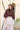 A woman stands outdoors in front of a beige wooden barn wall, wearing a dark brown wide-brim hat, a dark brown sheer long-sleeve blouse, a light blush pink tiered tulle skirt, and a patterned belt with a large gold buckle.Pliss_Tiered_Maxi_Skirt-FleaStyle