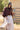 A woman wearing a brown ruffled blouse, a blush pleated tiered maxiskirt, a western belt with a heart-shaped buckle, and a beige cowboy hat, standing outdoors in front of a rustic barn-Pliss_Tiered_Maxi_Skirt-FleaStyle