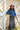 A woman wearing rust cowboy hat, a blue denim raw hemmed cropped jacket, a sheer lace long sleeve top, and a long dark flare olive skirt with silver studs, standing outdoors in front of a yellow wooden barn door-Suede_Western_Studded_Maxi_Skirt_-_Olive-FleaStyle