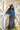 A woman wearing rust cowboy hat, a blue denim raw hemmed cropped jacket, a sheer lace long sleeve top, and a long dark flare olive skirt with silver studs, standing outdoors in front of a yellow wooden barn door-Suede_Western_Studded_Maxi_Skirt_-_Olive-FleaStyle