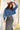 A woman wearing rust cowboy hat, a blue denim raw hemmed cropped jacket, a sheer lace long sleeve top, and a long dark flare olive skirt with silver studs, standing outdoors in front of a yellow wooden barn door-Suede_Western_Studded_Maxi_Skirt_-_Olive-FleaStyle