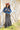 A woman wearing rust cowboy hat, a blue distressed denim raw hemmed cropped jacket, a sheer lace long sleeve top, and a long dark flared olive skirt with silver studs, standing outdoors in front of a yellow wooden barn door-Cropped_Distressed_Denim_Jacket-FleaStyle