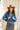 A woman wearing rust cowboy hat, a blue distressed denim raw hemmed cropped jacket, a sheer lace long sleeve top, and a long flared dark olive skirt with silver studs, standing outdoors in front of a yellow wooden barn door-Cropped_Distressed_Denim_Jacket-FleaStyle
