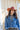 A woman wearing rust cowboy hat, a blue distressed denim raw hemmed cropped jacket, a sheer lace long sleeve top, and a long dark flared olive skirt with silver studs, standing outdoors in front of a yellow wooden barn door-Cropped_Distressed_Denim_Jacket-FleaStyle