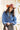 A woman wearing rust cowboy hat, a blue denim raw hemmed distressed cropped jacket, a sheer lace long sleeve top, and a long flared dark olive skirt with silver studs, standing outdoors in front of a yellow wooden barn door-Cropped_Distressed_Denim_Jacket-FleaStyle
