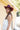 A woman standing against a yellow barn wearing a long ivory lace tiered halter dress, a maroon cowboy hat, and colorful rodeo silk scarf, and brown boots-Lace_Tiered_Halter_Maxi_Dress_-_Ivory-FleaStyle