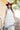 A woman standing against a yellow barn wearing a long ivory lace tiered halter dress, a maroon cowboy hat, and brown boots-Lace_Tiered_Halter_Maxi_Dress_-_Ivory-FleaStyle