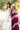 Two women standing outdoors in a sunlit field wearing bohemian-style dresses and wide-brim hats. One woman wears a long ivory lace tiered halter dress with a maroon hat, and the other wears a burgundy maxi dress with  pink lace trim and a flat brim hat-Lace_Tiered_Halter_Maxi_Dress_-_Ivory-FleaStyle.