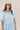 A woman wearing a light blue tiered short-sleeve ruffled shirt dress, a beige trucker cap with 'LOCAL' written on it, and brown cowboy boots, standing in front of a gold floral wallpaper background-Bubble_Sleeve_Button_Down_Babydoll_Dress_-_Baby_Blue-FleaStyle