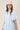 A woman wearing a light blue tiered short-sleeve ruffled shirt dress, a beige trucker cap with 'LOCAL' written on it, and brown cowboy boots, standing in front of a gold floral wallpaper background-Bubble_Sleeve_Button_Down_Babydoll_Dress_-_Baby_Blue-FleaStyle