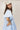 A woman wearing a light blue tiered short-sleeve ruffled shirt dress, a beige trucker cap with 'LOCAL' written on it, and brown cowboy boots, standing in front of a gold floral wallpaper background-Bubble_Sleeve_Button_Down_Babydoll_Dress_-_Baby_Blue-FleaStyle