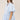 A woman wearing a light blue tiered short-sleeve ruffled shirt dress, a beige trucker cap with 'LOCAL' written on it, and brown cowboy boots, standing in front of a gold floral wallpaper background-Bubble_Sleeve_Button_Down_Babydoll_Dress_-_Baby_Blue-FleaStyle