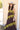 A woman wearing a sleeveless brown maxi dress featuring contrast olive lace detailing throughout for a rich, vintage-inspired feel paired with and a stone/beige cowboy hat, standing outdoors in front of a barn-Lace_Trim_Maxi_Dress_-_Brown_Chartreuse-FleaStyle