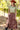 A woman wearing a  flowy maxi skirt featuring cascading asymmetrical tiers, raw-edge detailing and a dramatic side slit and an olive green graphic t-shirt stands outdoors in a wooded setting. She is also wearing a  rust cowboy hat and black boots-Asymmetrical_Tiered_Maxi_Skirt_-_Brown.