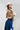 A woman wearing an ivory wide-flat brim hat, a beige suede- like fringed shrug, and a blue denim skirt against a light gray studio background.-Suede_Fringe_Shrug_-_Tan-FleaStyle