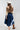 A woman shown from the back wearing an ivory wide-flat brim hat, a beige suede- like fringed shrug, beige button up shirt, blue denim skirt and cowboy boots against a light gray studio background.-Suede_Fringe_Shrug_-_Tan-FleaStyle