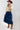 A woman wearing a tan button-up blouse with pearl embellished details, a maroon cowboy hat, and a dark blue denim midi skirt, photographed against a light gray studio background-Pearl_Embellished_Button_Down_Blouse_-_Tan-FleaStyle