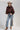 A woman wearing a brown satin cape-style blouse with a large bow at the neckline, a beige cowboy hat, and embroidered floral blue jeans, photographed against a plain light gray studio background-Exaggerated_Bow_Blouse_-_Rust-FleaStyle