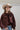 A woman wearing a brown satin cape-style blouse with a large bow at the neckline, a beige cowboy hat, and embroidered floral blue jeans, photographed against a plain light gray studio background-Exaggerated_Bow_Blouse_-_Rust-FleaStyle