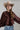 A woman wearing a brown satin cape-style blouse with a large bow at the neckline, a beige cowboy hat, and embroidered floral blue jeans, photographed against a plain light gray studio background-Exaggerated_Bow_Blouse_-_Rust-FleaStyle
