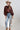 A woman wearing a brown satin cape-style blouse with a large bow at the neckline, a beige cowboy hat, and embroidered floral blue jeans, photographed against a plain light gray studio background-Exaggerated_Bow_Blouse_-_Rust-FleaStyle