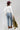 A woman wearing a white off-the-shoulder long-sleeve crop top, embroidered floral blue jeans and a  maroon cowboy hat, and holding an olive green faux leather jacket, against a light gray studio background-Floral_Embroidered_High_Waist_Flare_Jeans-FleaStyle.