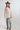 A woman wearing a maroon wide-brim cowboy hat and a sheer, pleated light blush flowy cape blouse with a large tie bow at the collar, paired with blue jeans, set against a plain light gray background-Pliss_Tie-Neck_Cape_Top-FleaStyle.