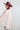 A woman wearing a maroon wide-brim cowboy hat and a sheer, pleated light blush flowy cape blouse with a large tie bow at the collar, paired with blue jeans, set against a plain light gray background-Pliss_Tie-Neck_Cape_Top-FleaStyle.