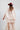 A woman wearing a maroon wide-brim cowboy hat and a sheer, pleated light beige flowy cape blouse with a large tie bow at the collar, paired with a matching flowy ruffle pleated maxi skirt, set against a plain light gray background-Pliss_Tiered_Maxi_Skirt-FleaStyle