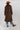 A woman wearing a long,  brown faux fur coat with a horse motif lining, paired with a rust-colored cowboy hat, a maroon tiger patterned top, and blue jeans against a gray background-Longline_Faux_Fur_Jacket_-_Brown-FleaStyle
