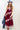 A woman wearing a  maxi dress in deep burgundy with light pink floral lace trim, styled with a dusty blue cowboy hat and holding a blue denim jacket against a plain white studio background.-Lace_Trim_Maxi_Dress_-_Blush_Maroon-FleaStyle