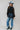 A woman wearing a sheer black lace top in a flowy silhouette, scalloped detailing and an oversized bow and a high ruffled collar, paired with dusty blue cowboy hat and blue jeans, against a plain light gray background-Sheer Lace Tie Neck Blouse - Black