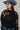 A woman wearing a sheer black lace top in a flowy silhouette, scalloped detailing and an oversized bow and a high ruffled collar, paired with dusty blue cowboy hat and blue jeans, against a plain light gray background-Sheer Lace Tie Neck Blouse - Black