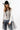 A woman wearing a western-style outfit featuring a white long-sleeve button-up shirt, a sheer black mesh overlay, featuring a relaxed, sleeveless fit and a flowy tulle hem, blue jeans, a black cowboy hat, and black ankle boots, against a plain white studio background-Sleeveless_Sheer_Mesh_Overlay_-_Black