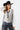 A woman wearing a western-style outfit featuring a white long-sleeve button-up shirt, a sheer black mesh overlay, featuring a relaxed, sleeveless fit and a flowy tulle hem, blue jeans, a black cowboy hat, and black ankle boots, against a plain white studio background-Sleeveless_Sheer_Mesh_Overlay_-_Black-FleaStyle