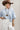 A model wearing a light blue knit short-sleeve top, modern barrel-leg silhouette, adjustable drawstring waist and utility-inspired seaming for a relaxed yet elevated fit, a brown straw cowboy hat, and blue boots--Drawstring_Barrel_Jogger_Pants_-_Ivory-FleaStyle