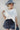 A woman wearing a relaxed, boxy fit cropped tee with a baby blue heart graphic that reads 'Howdy Wild & Free' , paired with dark paisley patterened denim shorts and a wide-brimmed dusty blue cowboy hat-Howdy_Heart_Western_Motif_Graphic_T-Shirt-FleaStyle