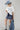 A woman wearing a relaxed, boxy fit cropped tee with a baby blue heart graphic that reads 'Howdy Wild & Free' , paired with dark paisley patterened denim shorts and a wide-brimmed dusty blue cowboy hat-Howdy_Heart_Western_Motif_Graphic_T-Shirt-FleaStyle