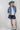 A woman wearing a western-style outfit consisting of a light blue relaxed cropped denim shirt-jacket, dark blue denim shorts with a paisley pattern, a white graphic t-shirt, a dusty blue cowboy hat, and silver cowboy boots-
