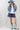 A woman wearing a western-style outfit consisting of a light blue relaxed cropped denim shirt-jacket, dark blue denim shorts with a paisley pattern, a white graphic t-shirt, a dusty blue cowboy hat, and silver cowboy boots-
