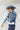 A woman wearing a western-style outfit consisting of a light blue relaxed cropped denim shirt-jacket, dark blue denim shorts with a paisley pattern, a white graphic t-shirt, a dusty blue cowboy hat, and silver cowboy boots-