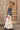 A woman modeling a strapless blue corset-style top with gold-tone clasps, a long satin maxi skirt with a colorful printed  horse design, paired with a beige cowboy hat and dark blue boots, against studio background-