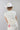 A woman wearing a cream-colored drawstring top with an easy relaxed fit ,adjustable side ruching and ties, a matching cream wide-brim hat, layered gold necklaces, and a white skirt with colorful horse prints, posing against a light gray studio background-Ruched_Drawstring_Top_-_Ivory-FleaStyle