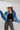 A model wearing a  cropped denim jacket featuring an oversized dolman-sleeve silhouette with unexpected asymmetrical lace detailing , paired with leopard-print pants, an ivory cowboy hat, and shiny silver cowboy boots, photographed against a light gray studio background--Lace_Trim_Dolman_Denim_Jacket-FleaStyle