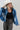 A model wearing a  cropped denim jacket featuring an oversized dolman-sleeve silhouette with unexpected asymmetrical lace detailing , paired with leopard-print pants, an ivory cowboy hat, and shiny silver cowboy boots, photographed against a light gray studio background--Lace_Trim_Dolman_Denim_Jacket-FleaStyle