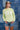 A woman wearing a lime green ribbed long-sleeve crewneck sweater with 'TEXAS' printed in white block letters across the chest, paired with denim shorts and a camouflage baseball cap, standing against a blue denim patchwork backdrop-Fleastyle