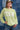 A woman wearing a lime green ribbed long-sleeve crewneck shirt with 'TEXAS' printed in white block letters across the chest, paired with denim shorts and a camouflage baseball cap, standing against a blue denim patchwork backdrop-Fleastyle